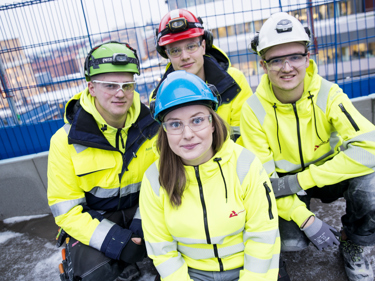 Fyra personer i varselvästar sitter på en byggarbetsplats och tittar rätt in i kameran