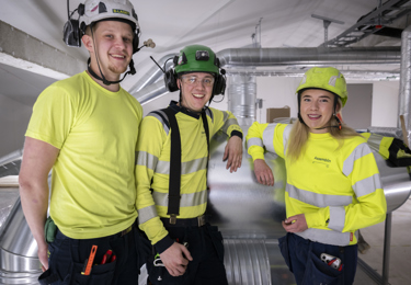 Två leende killar och en tjej i gula varselkläder och med hjälmar på huvudet